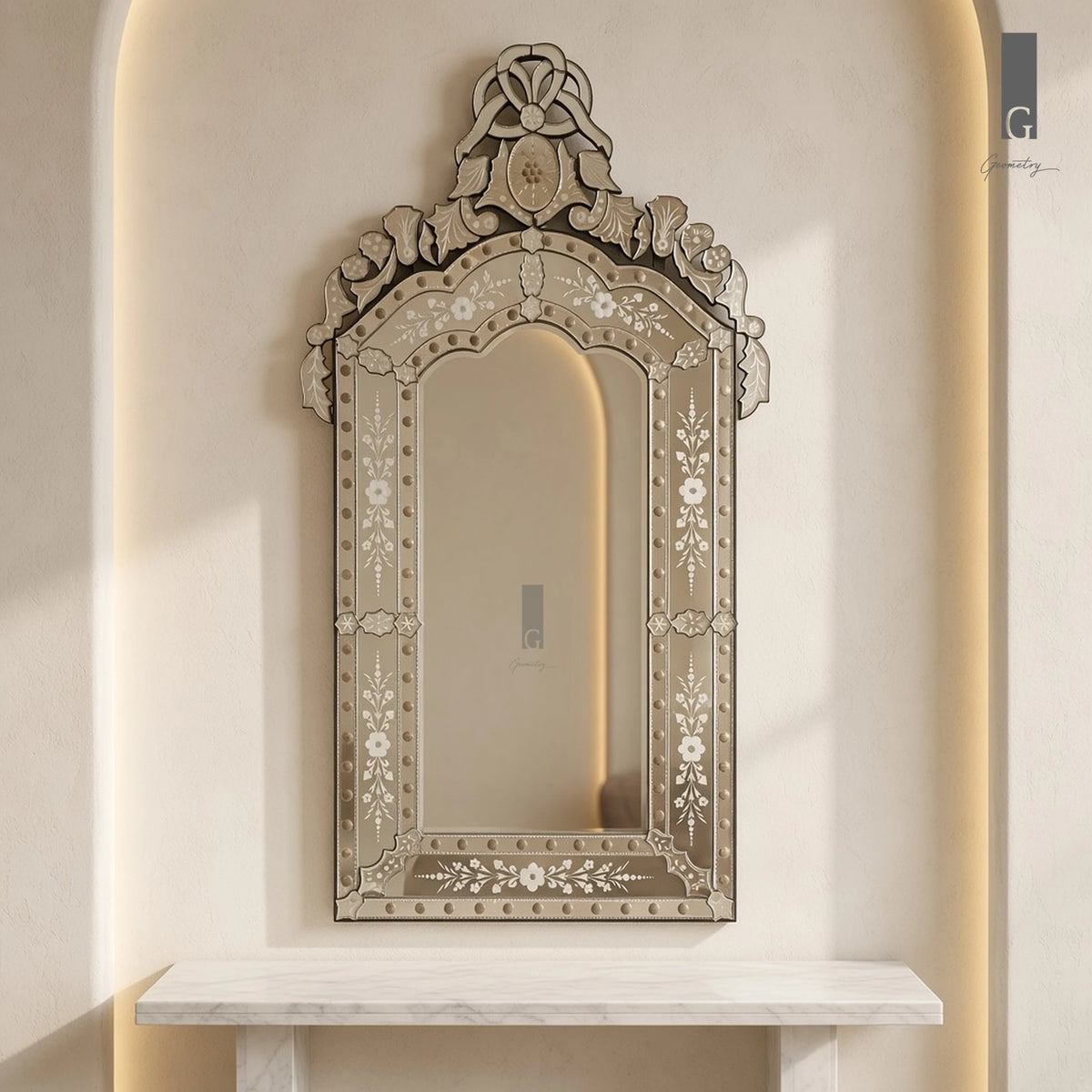 Geometry Designs Venetian Mirror in etched silver glass with studded frame and crest, shown above a white marble console in a warm arched niche.